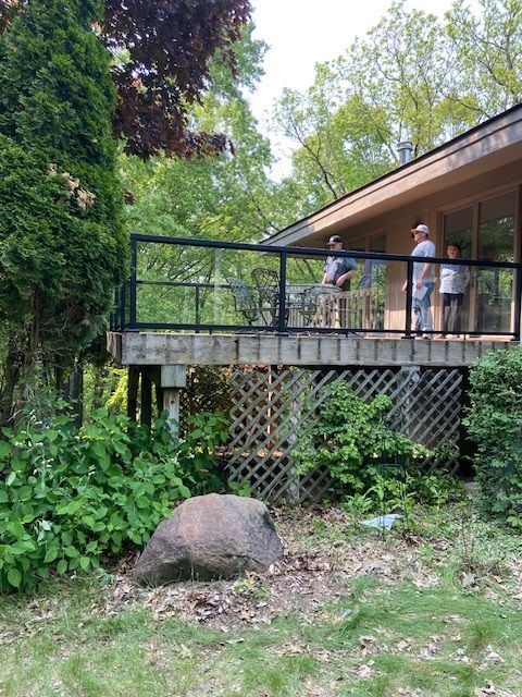 A raised deck with people on it. Glass railing, green trees surround. Large rock in the foreground.