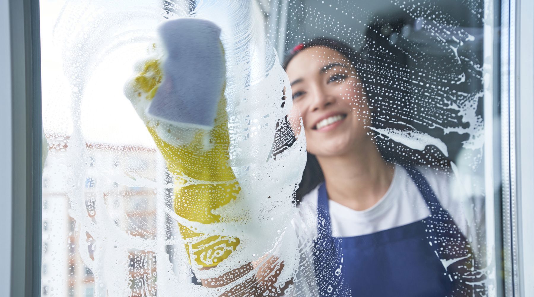 Woman wearing a blue apron and yellow gloves, smiles while cleaning a window with soapy water.