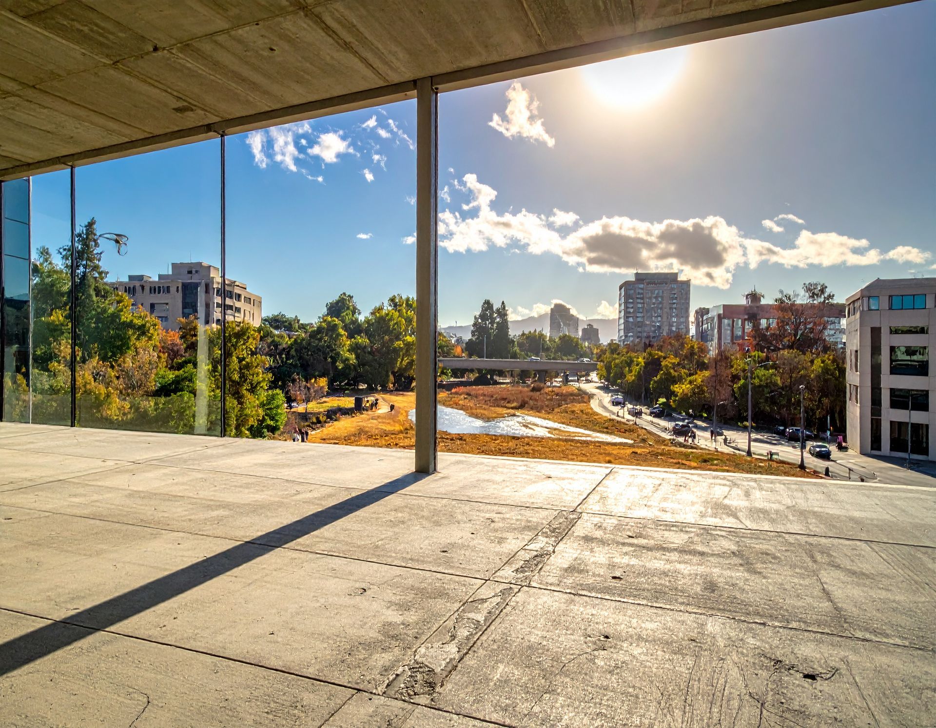 View from a modern, concrete interior overlooking a sunny city landscape with trees and buildings.