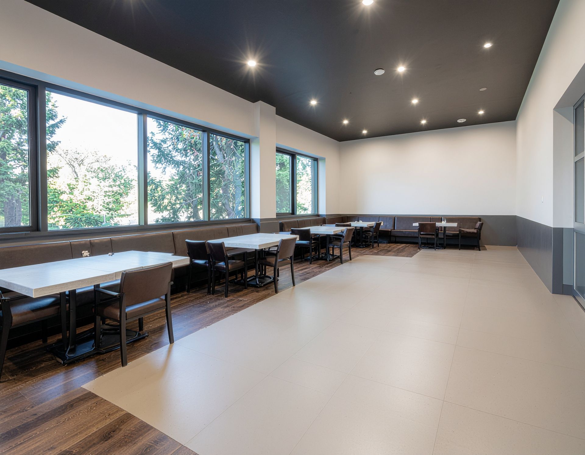 Spacious dining area with tables, chairs, and large windows overlooking trees. Gray and white walls, dark ceiling.