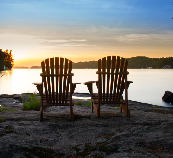 Two wooden Adirondack chairs face a lake at sunset.