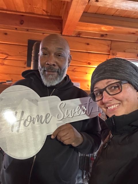 Man and woman holding a "Home Sweet" key-shaped sign, smiling. Interior setting with wooden beams.