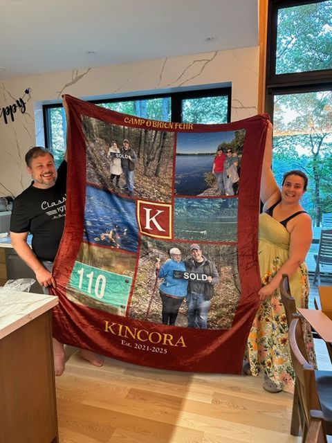 Couple holding a custom burgundy blanket with photos and text. "Kincora, Est. 1973-2023" is printed on the blanket.