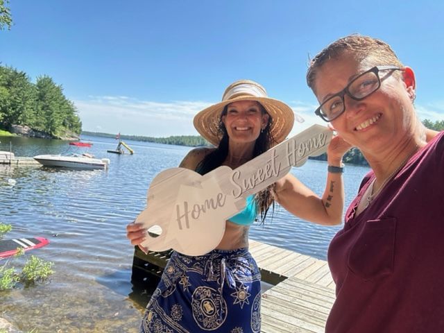 Two women on a dock by a lake, holding a large key-shaped sign that reads "Home Sweet Home".