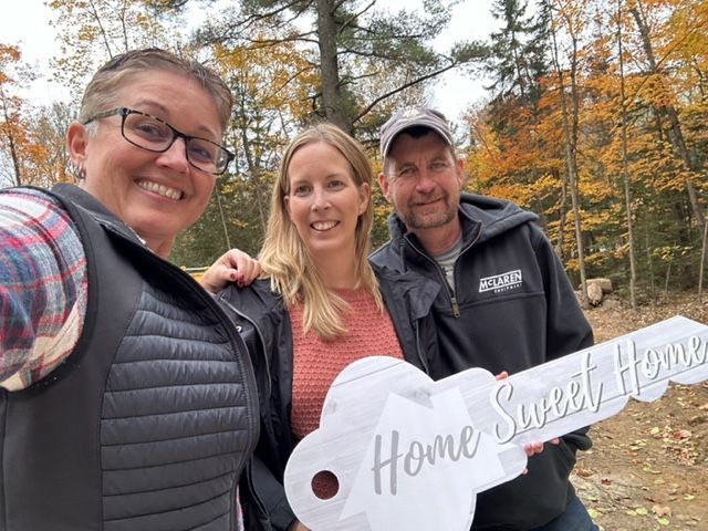 Three people holding a "Home Sweet Home" key-shaped sign, smiling. Autumn trees in the background.