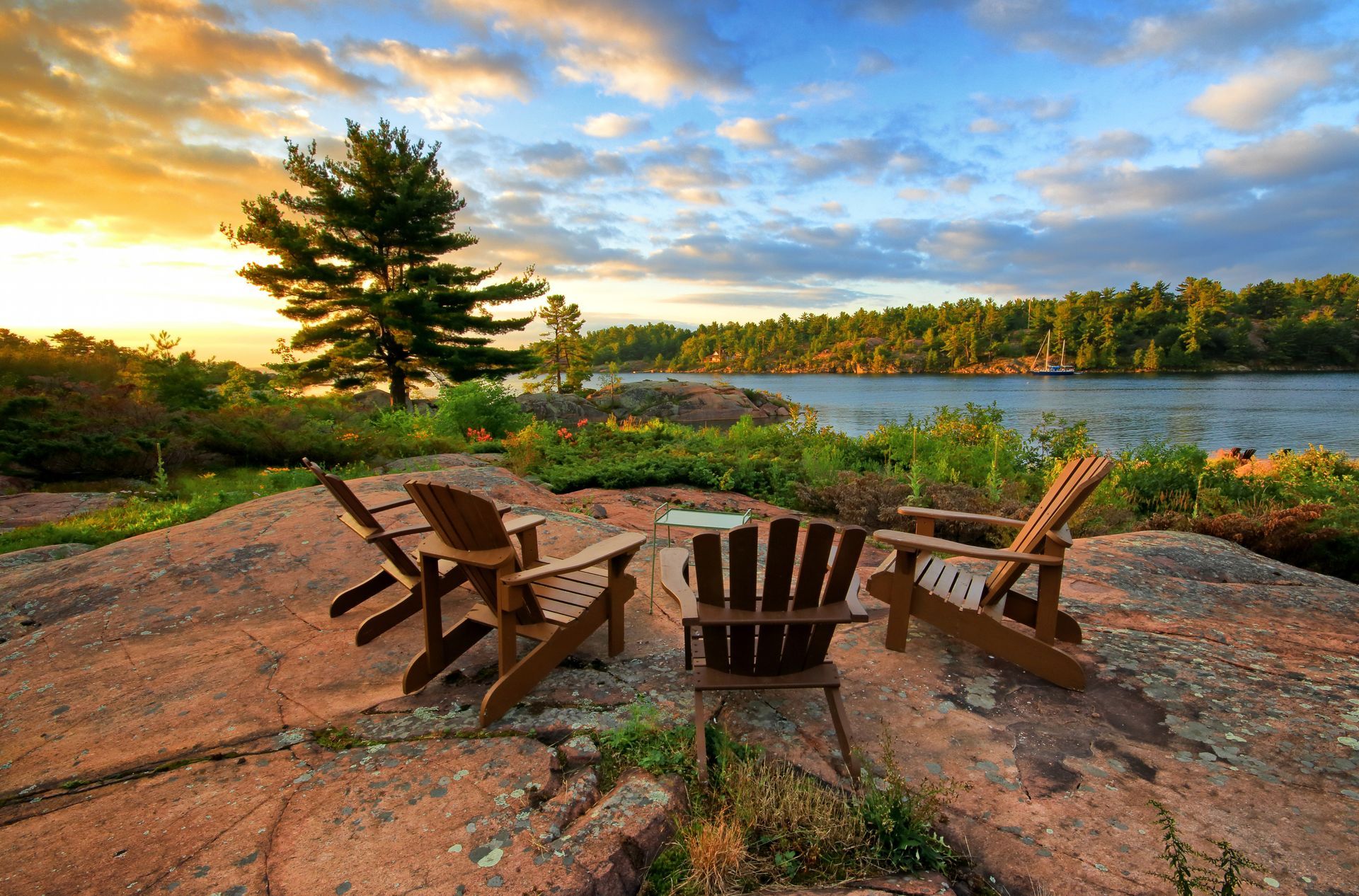 Wooden chairs around a small fire pit on a rocky shore overlooking a lake at sunset.
