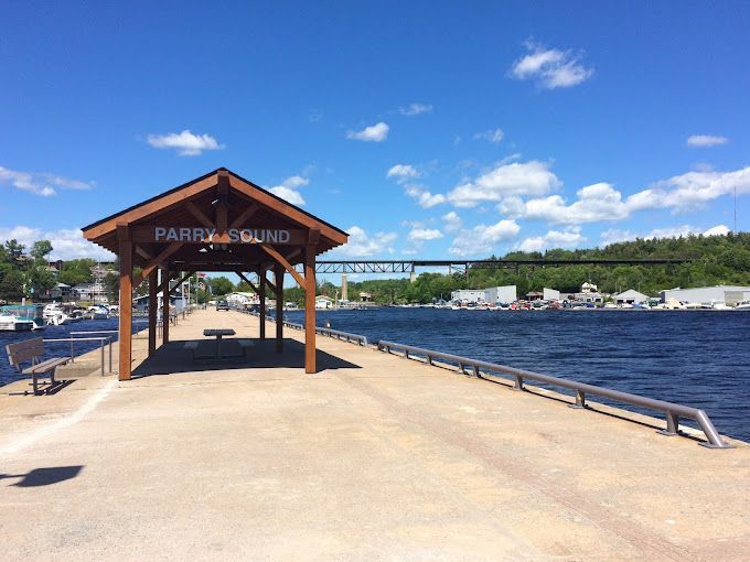 A pier in Parry Sound, Canada, with a covered shelter. A bridge and buildings are in the background under a blue sky.
