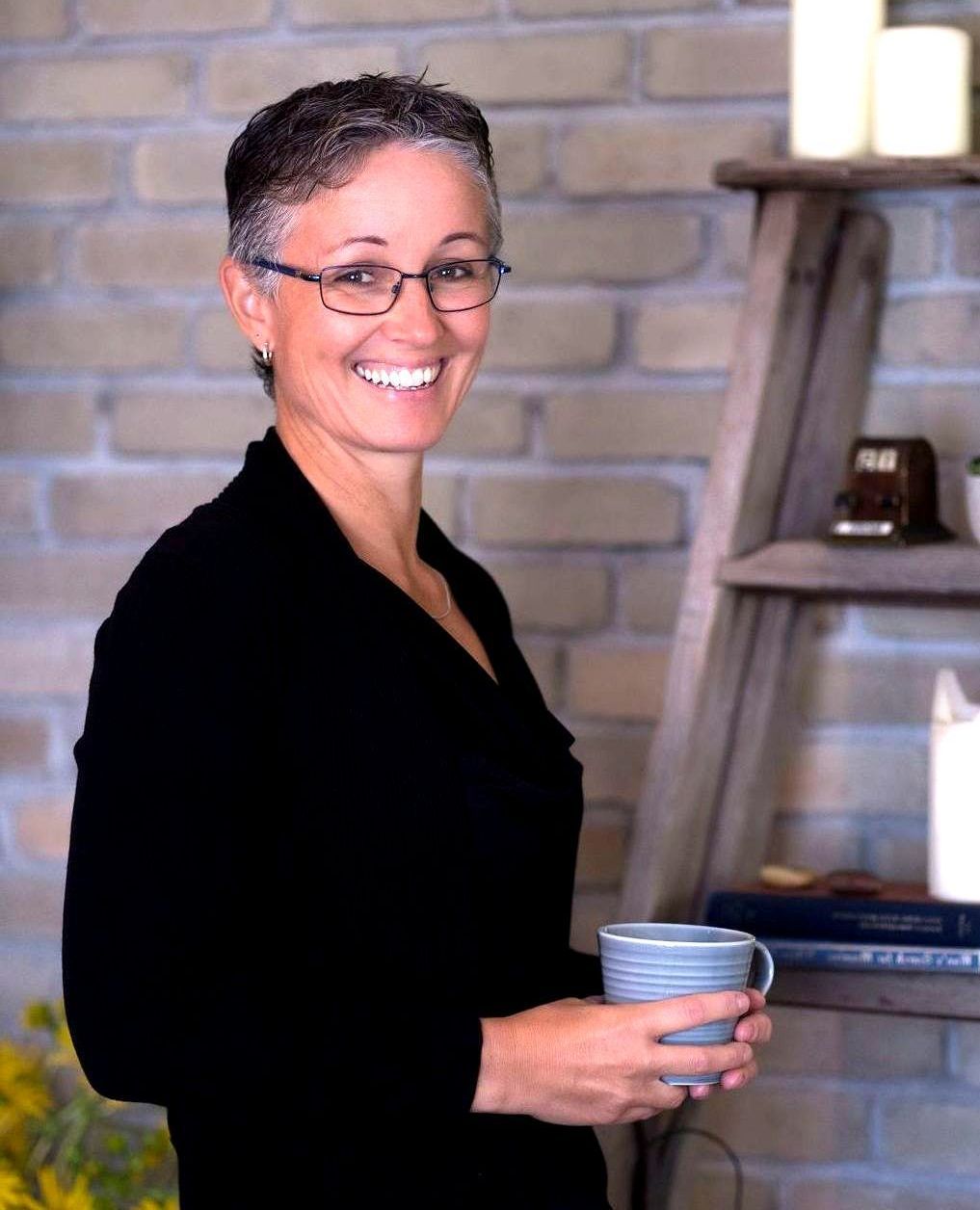 Woman with short, gray hair and glasses smiles, holding a mug. Leaning on a wooden ladder with candles and books, brick wall background.