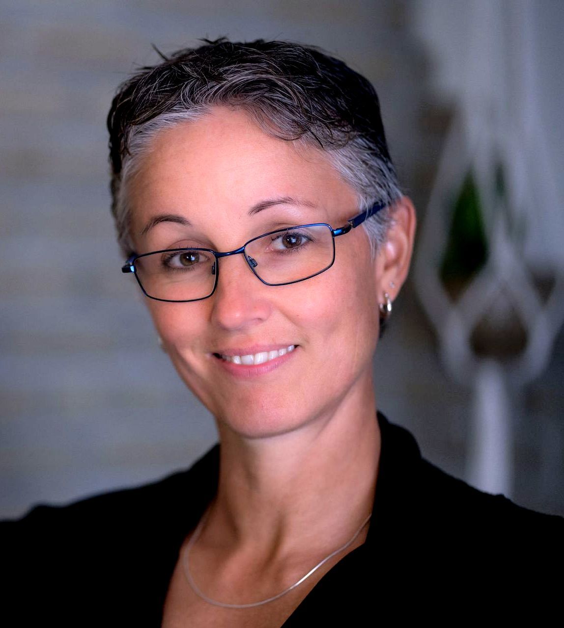 Woman with short, graying hair and glasses smiles, wearing black blazer.