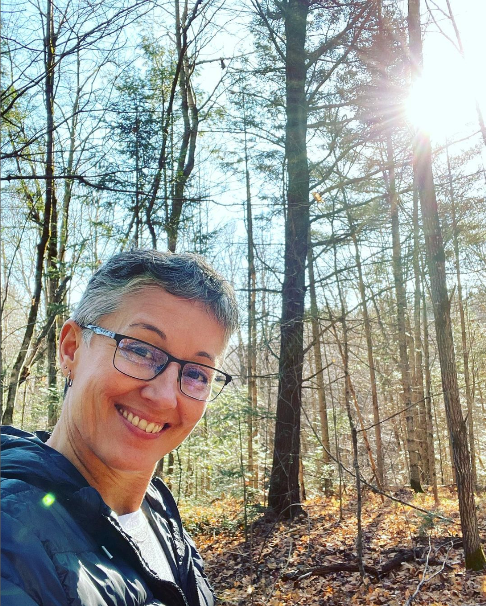 Woman smiling, wearing glasses, in a sunny forest with tall trees.