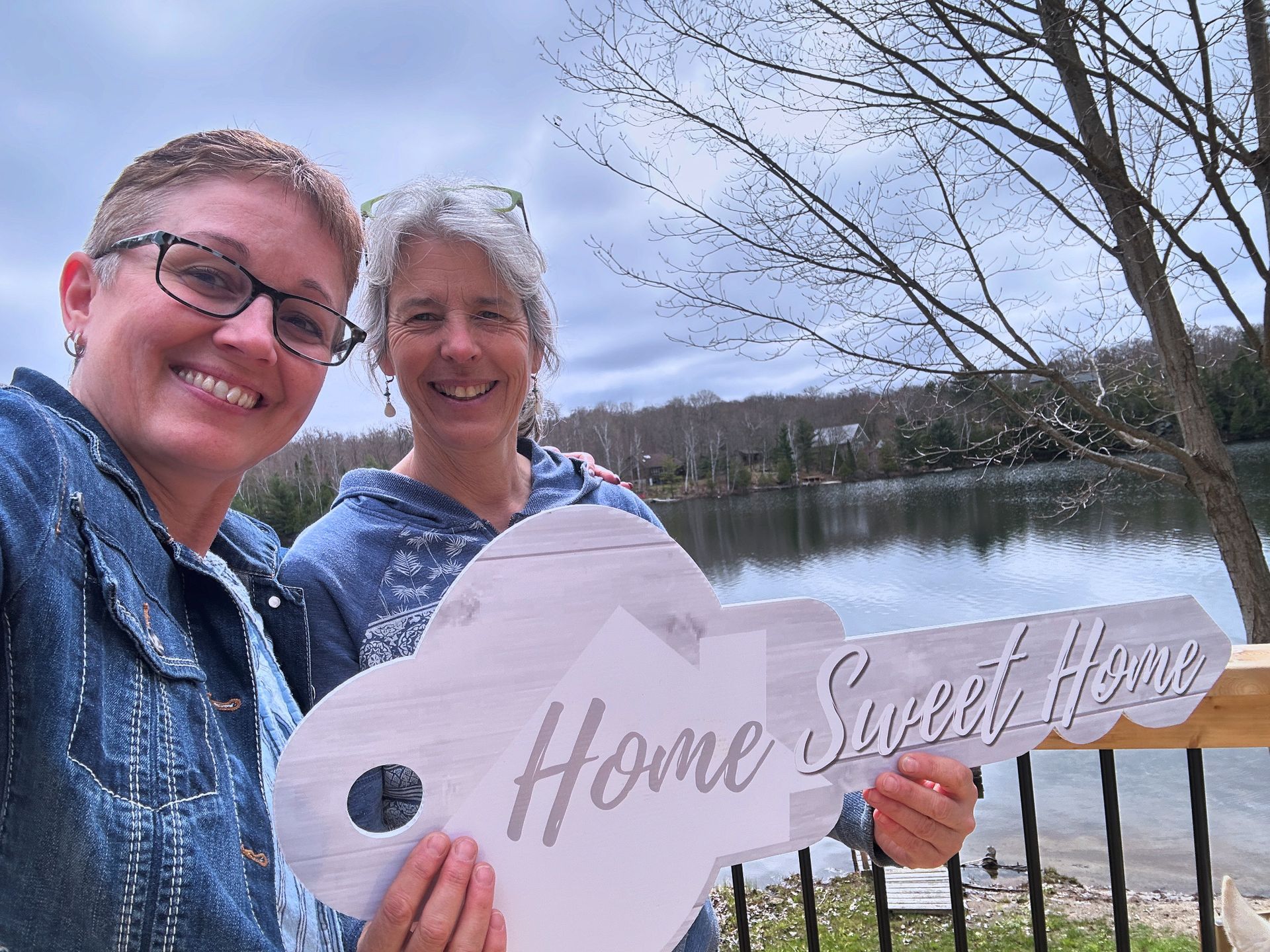 Two women holding a "Home Sweet Home" key-shaped sign, smiling by a lake.