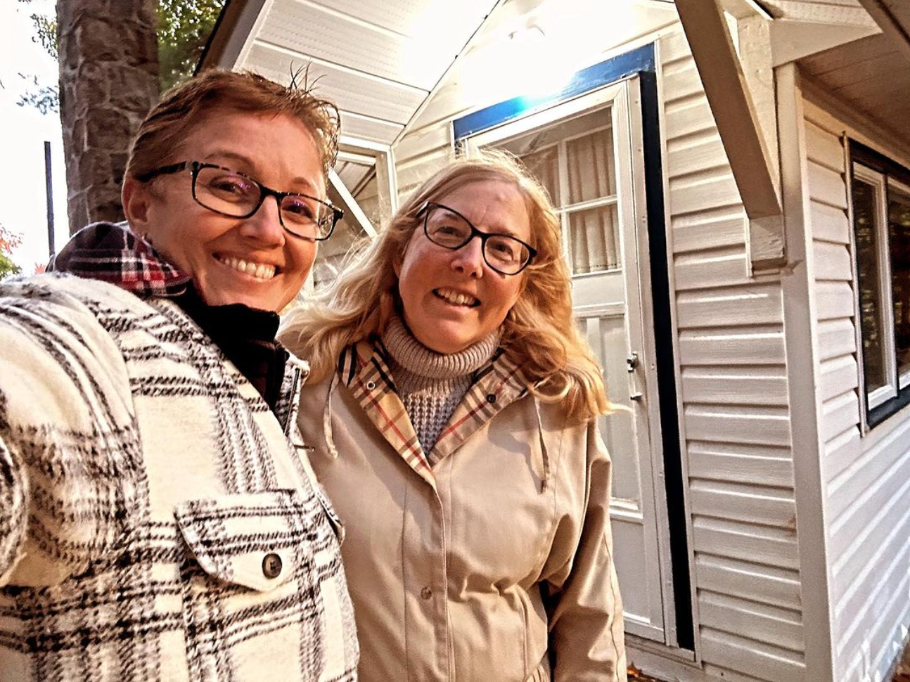 Two people smiling in front of a white building. One in a plaid shirt, the other in a tan coat.
