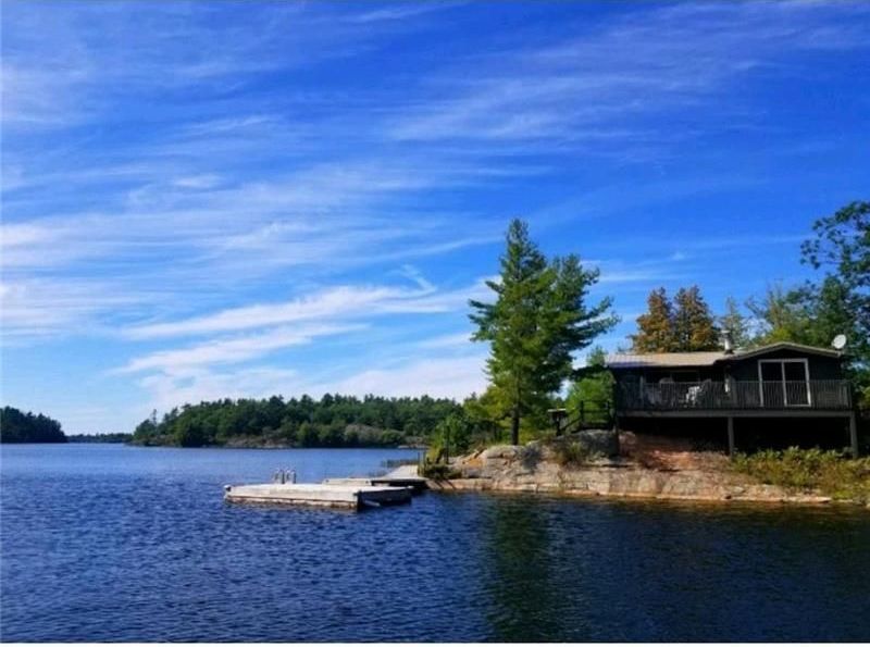 Lakeside cabin on a rocky shore under a bright blue sky with wispy clouds; a dock extends into the dark water.