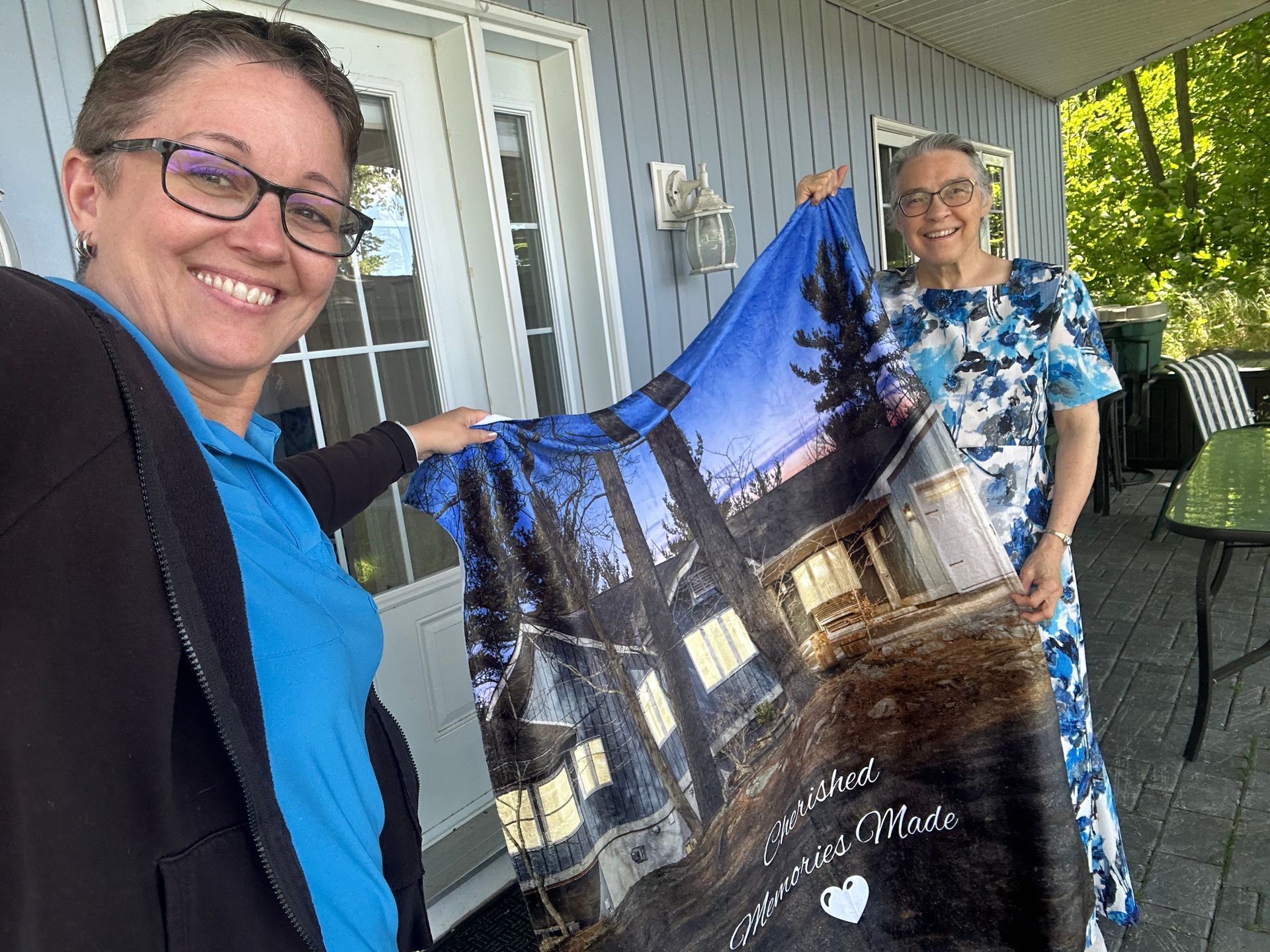 Two women smiling, holding a blanket with a house photo. Outdoors on porch.