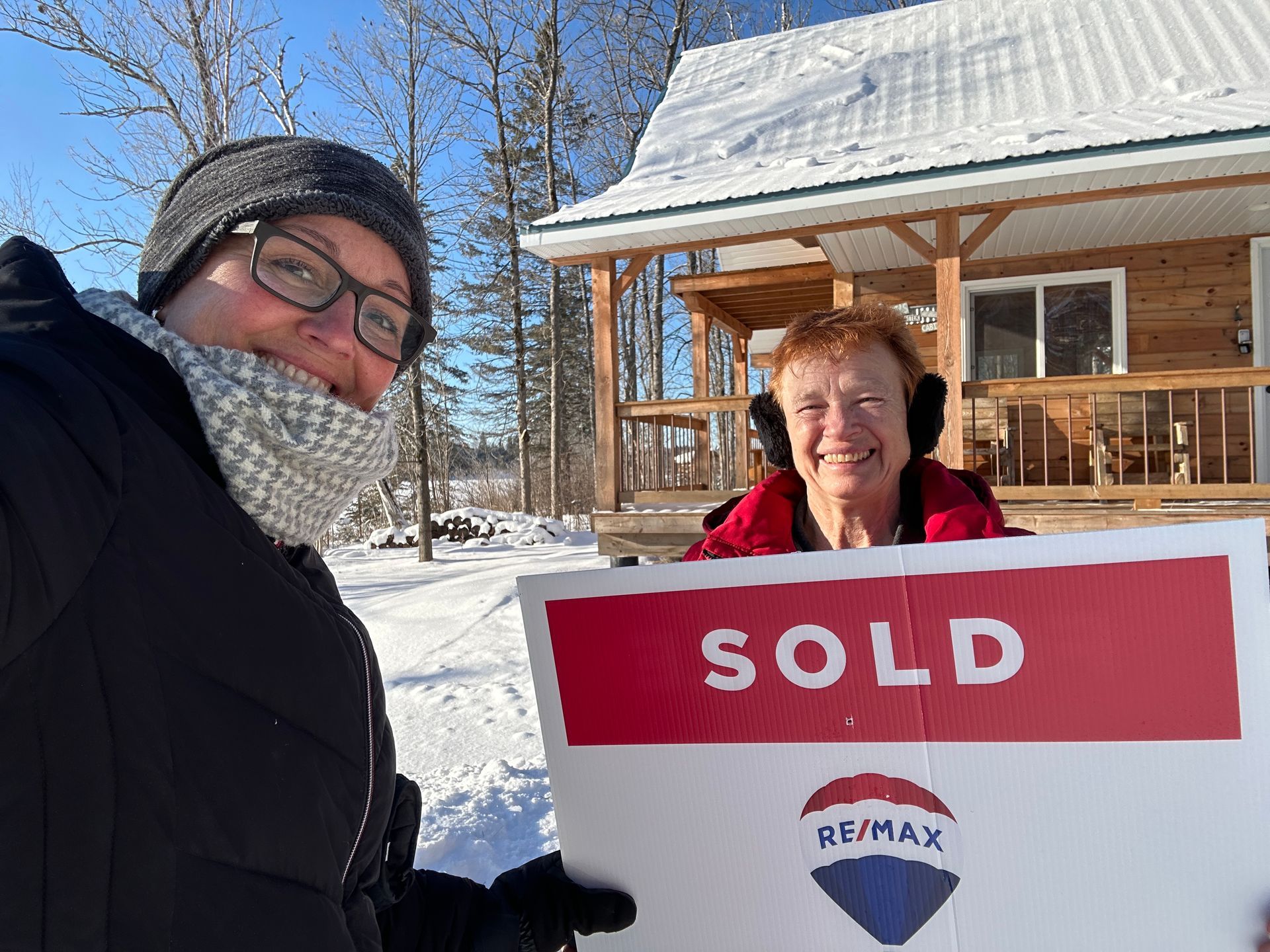 Two people holding "SOLD" sign in front of a snow-covered house; sunny day.