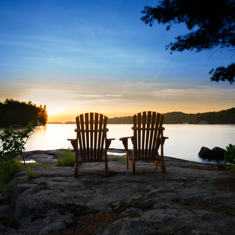 Two Adirondack chairs face a lake at sunset.
