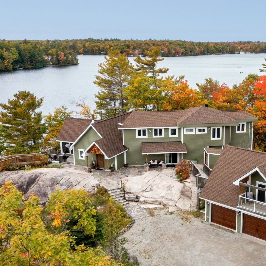 Green house on a rocky outcrop overlooking a lake with fall foliage. Brown roofs and white trim.