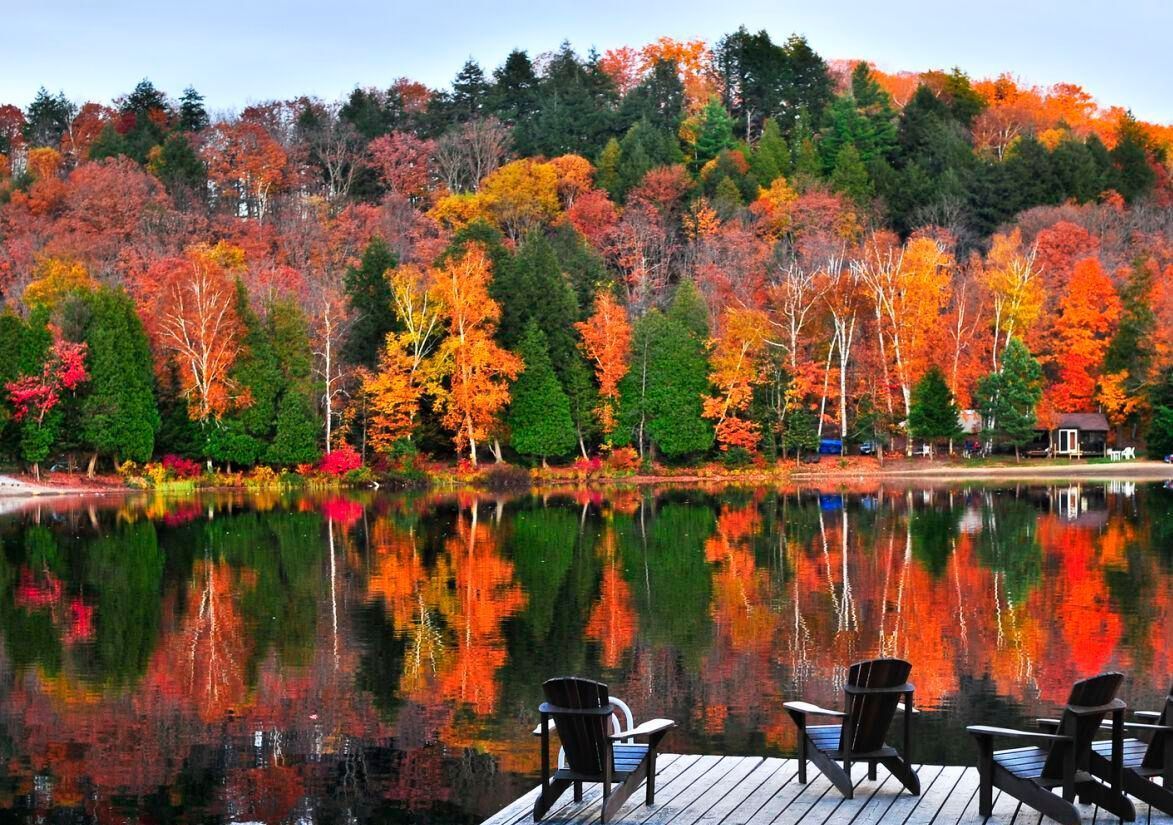 Autumn lake scene; vibrant red, orange, and green trees reflect in the water, with wooden chairs on a dock.