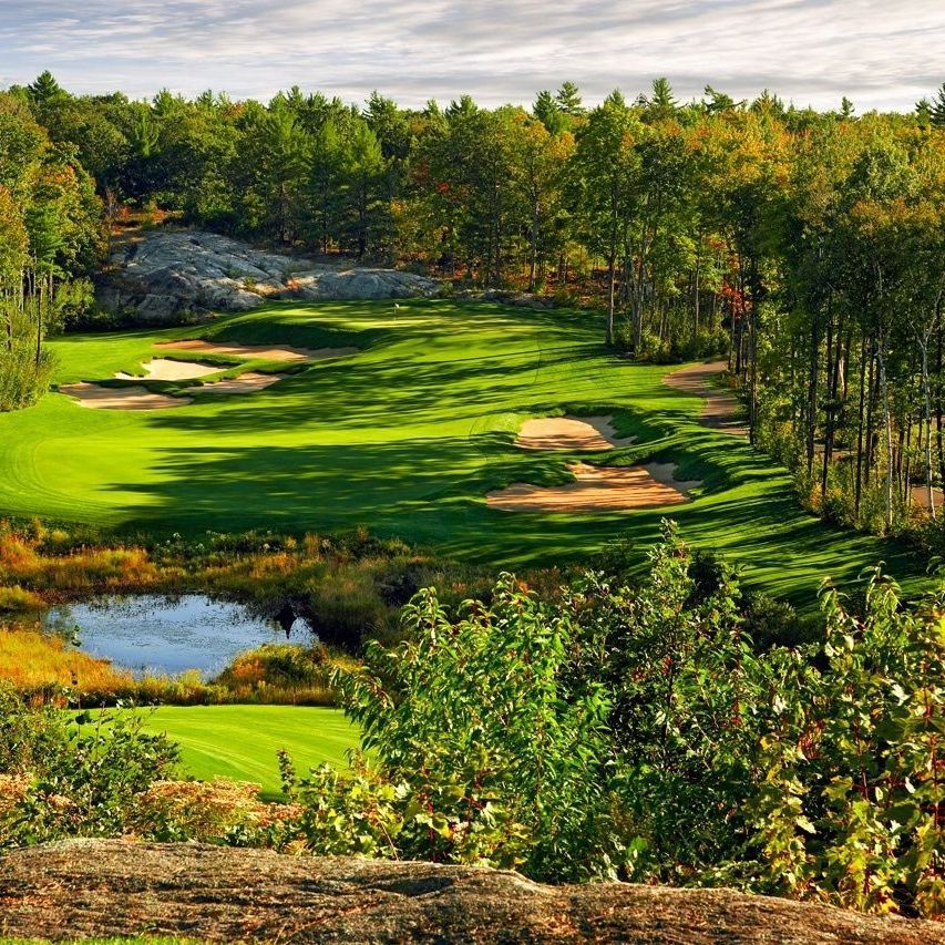 Green golf course with sand traps, a pond, and trees under a cloudy sky.