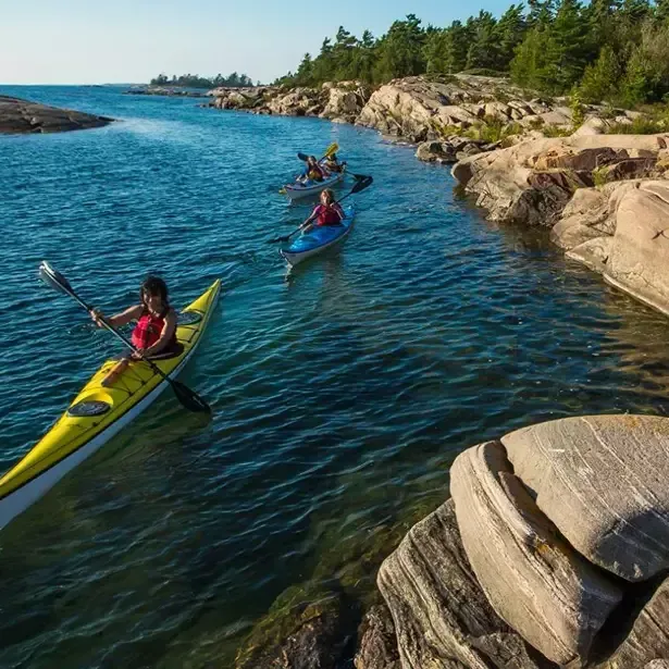 Kayakers paddle along a rocky shoreline, blue water, sunny day.