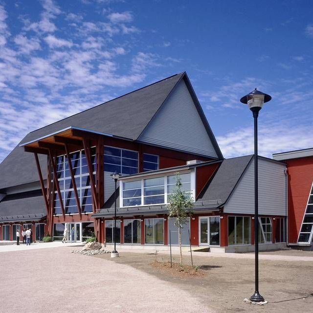 Exterior view of a modern red and gray building with a large roof and glass windows under a blue sky.