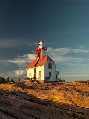 White lighthouse with red roof on rocky shore, lit by golden sunlight. Blue sky.