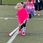 Child in bright pink jacket playing field hockey, guiding a ball on turf with a stick.