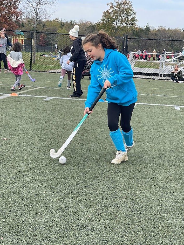 Child in blue hoodie playing field hockey on an outdoor turf field, with other players in the background.
