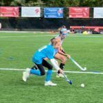 Two field hockey players in blue and tan sprint after the ball on a grassy field.
