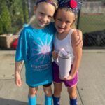 Two children standing outdoors, smiling and posing side by side in bright athletic clothes.