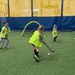 Children playing indoor hockey on turf, one dribbling a ball near a small goal.