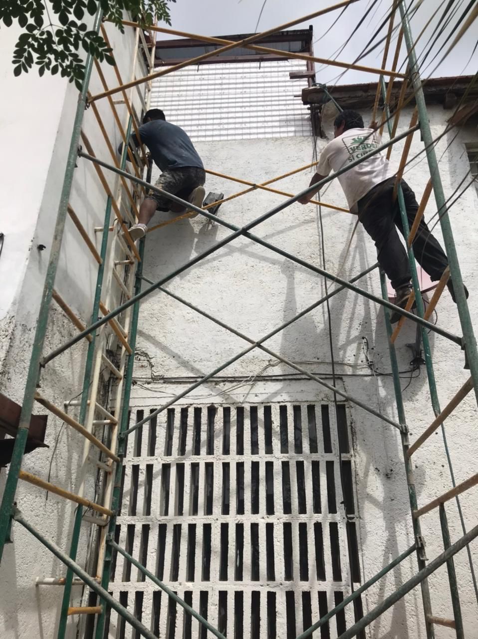 Dos trabajadores sobre andamios reparan el exterior blanco de un edificio cerca de una rejilla de ventilación negra, en un día soleado.