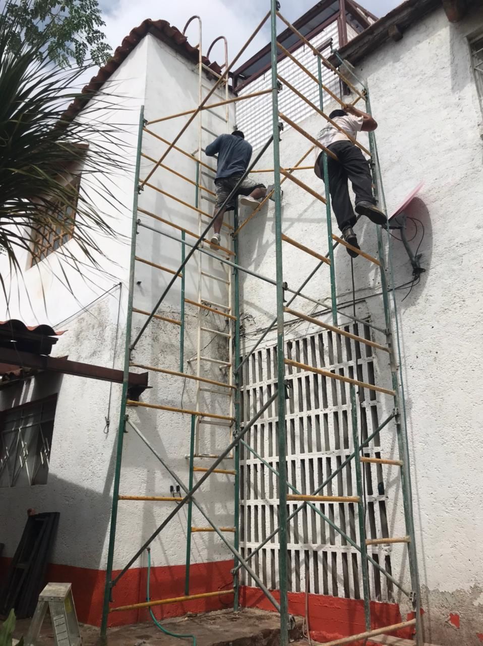 Dos personas trabajando en el exterior de un edificio, utilizando un andamio metálico alto. Pared de estuco blanco.