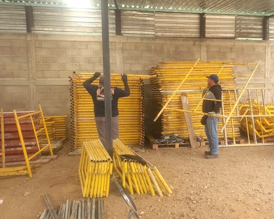 Dos trabajadores clasificando andamios amarillos en un almacén. Materiales de construcción apilados a gran altura.