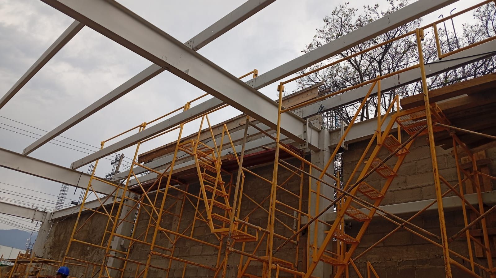 Obra en construcción con vigas de acero, andamios y paredes color canela bajo un cielo nublado.