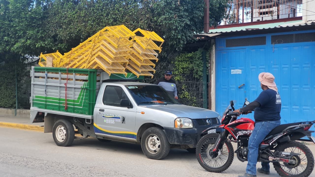 Un camión cargado de sillas metálicas amarillas está estacionado cerca de una puerta azul. Una persona está sentada en una motocicleta roja cerca.