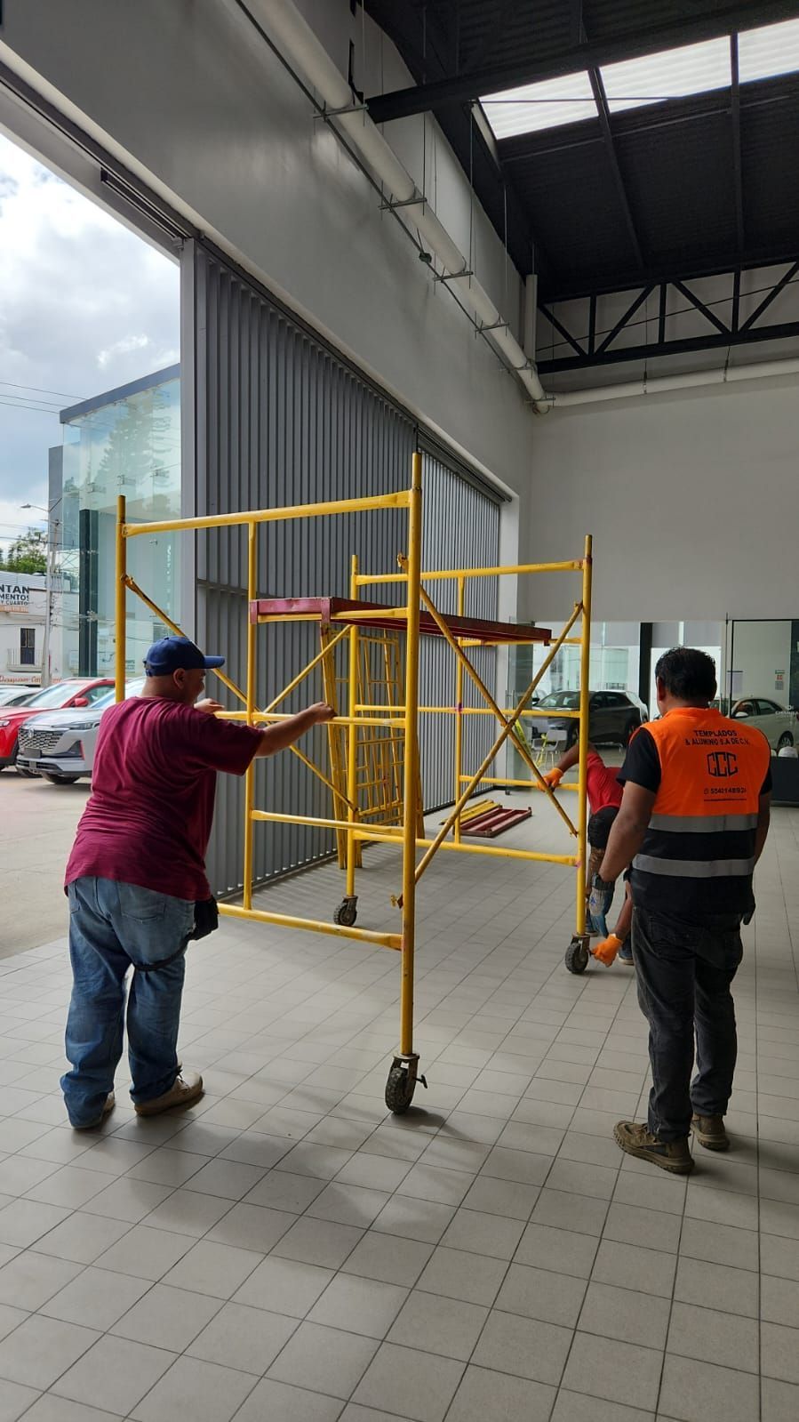 Dos trabajadores trasladan un andamio móvil amarillo al interior de un gran edificio con la puerta abierta.