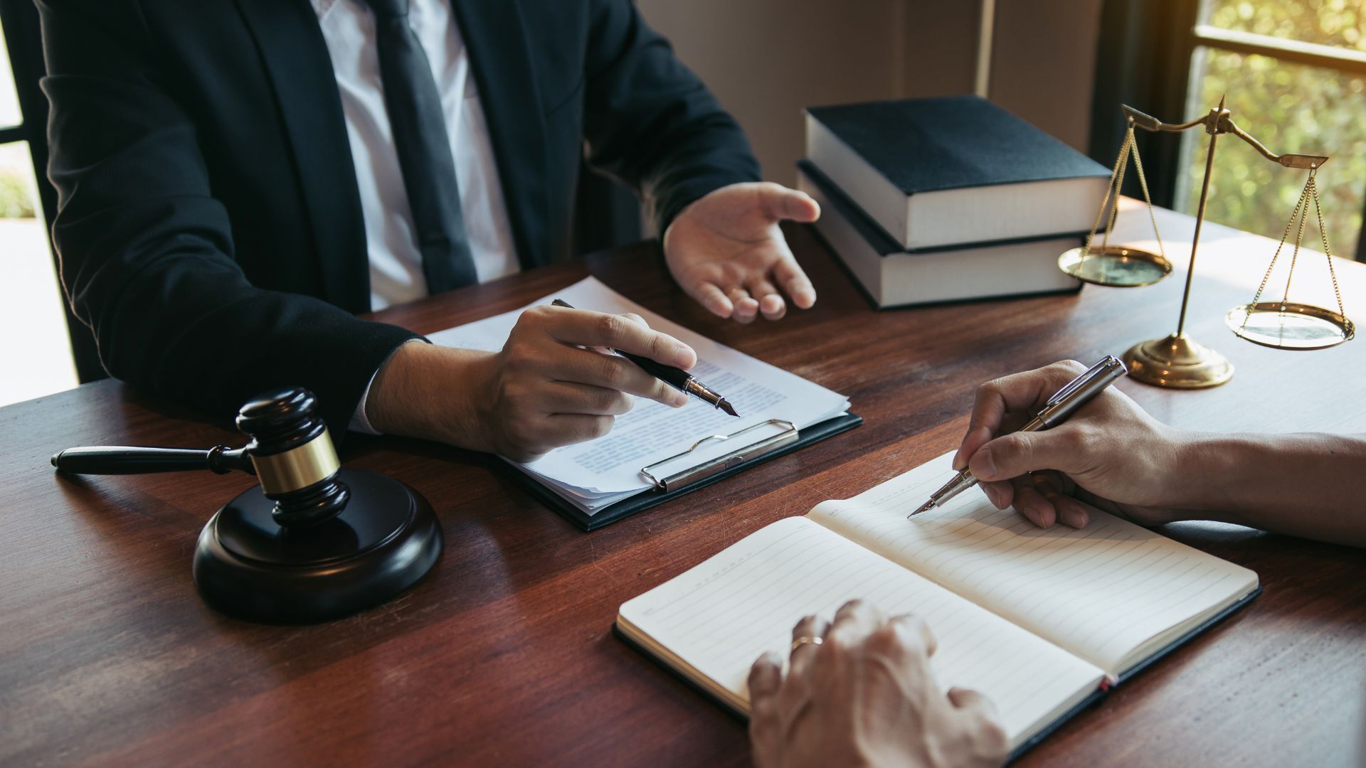 Lawyer in suit gestures, reviewing paperwork with client; gavel, scales, and books on table.