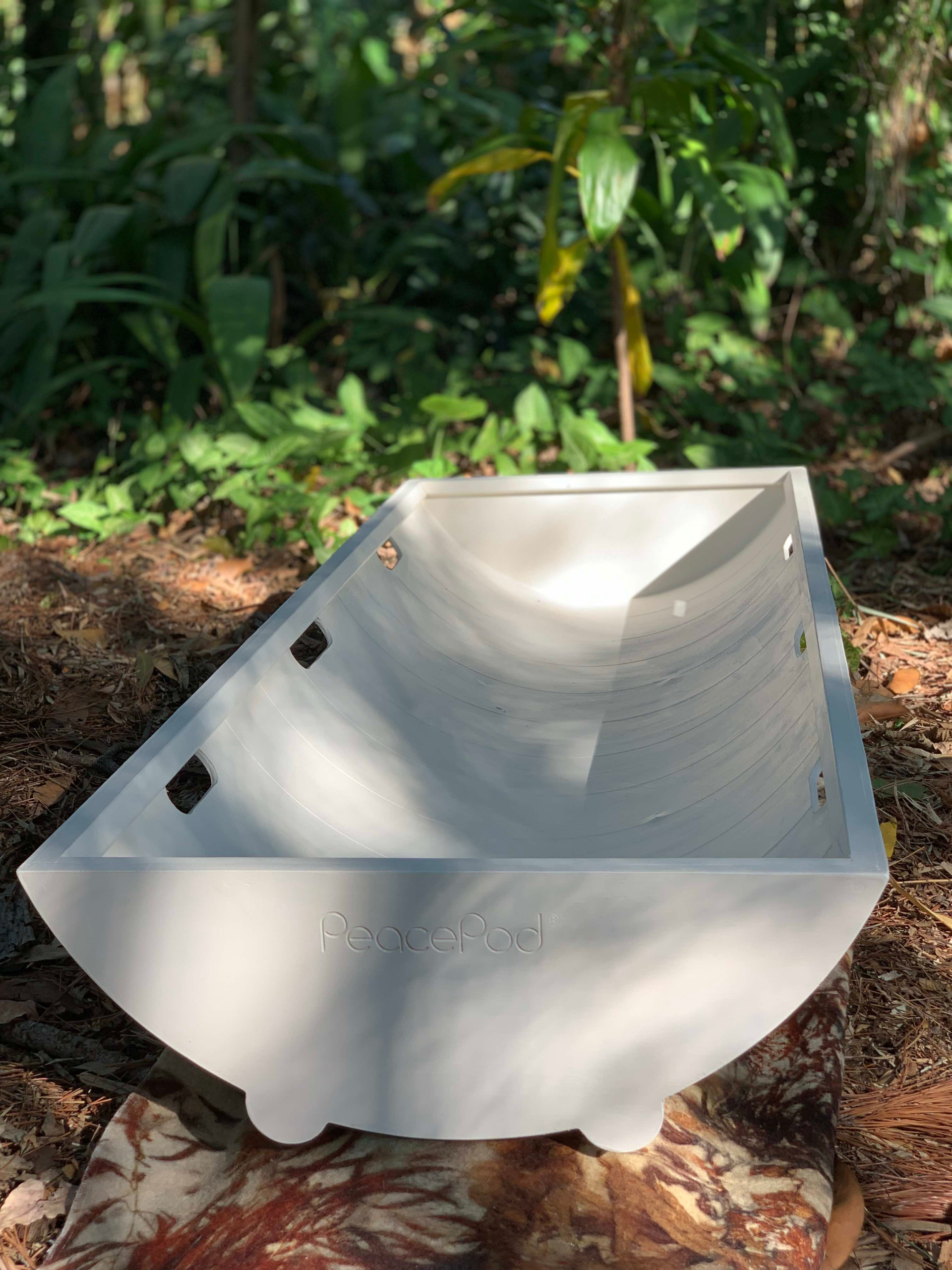 A white PeacePod shroud bearer tray is sitting on top of a pile of leaves in the woods.