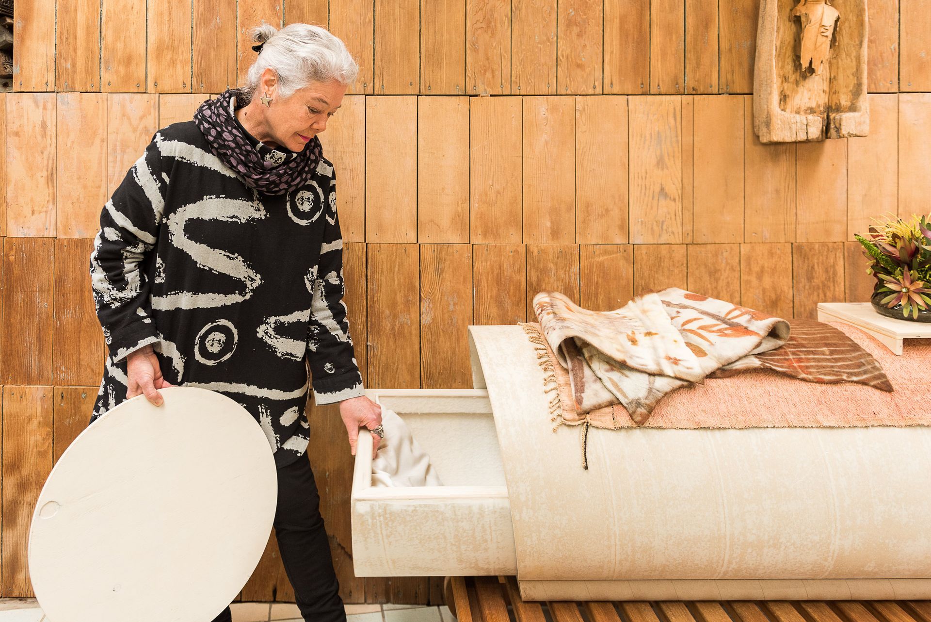 A woman is standing in front of a wooden wall next to a PeacePod casket holding the end of the casket.