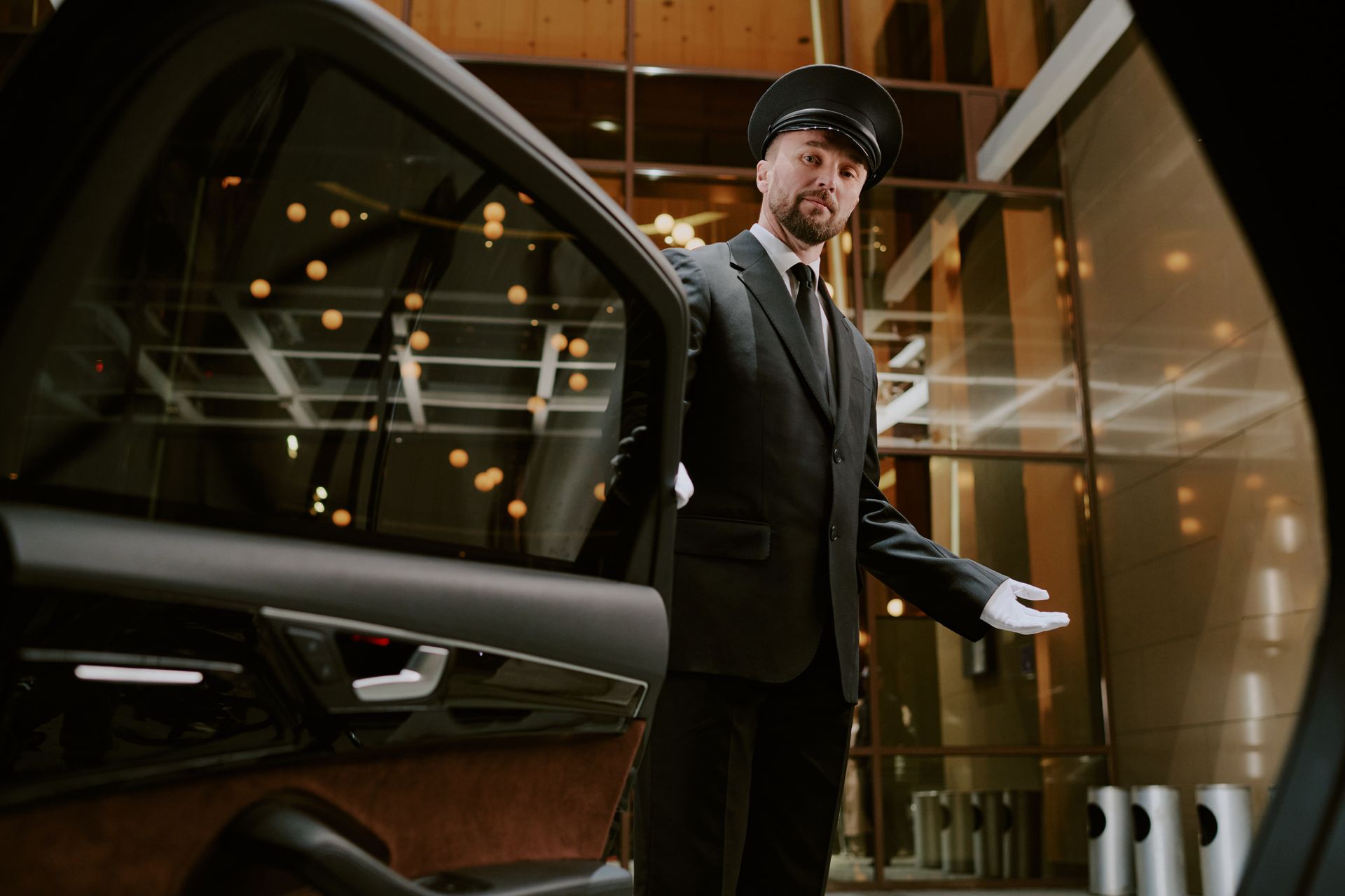 Man in a suit holding a car door open in a modern showroom, with warm lights and glass walls.
