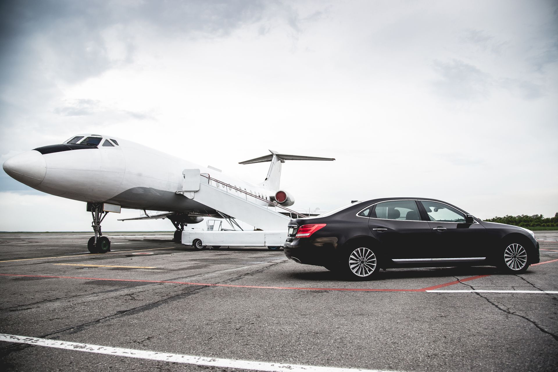 Private jet beside a black sedan on an airport tarmac under an overcast sky.