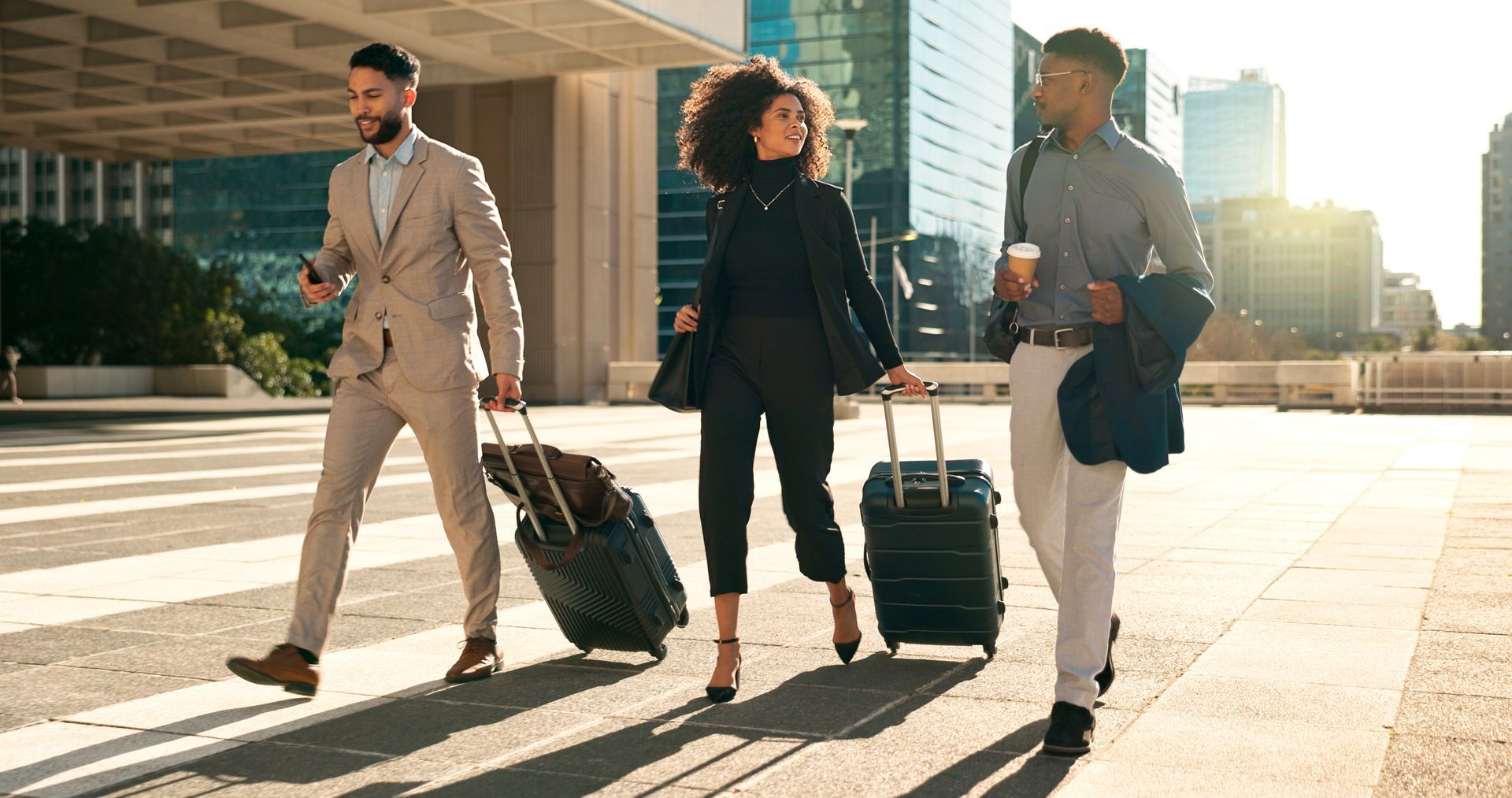 Three business travelers walk with luggage outside a modern office building in bright sunlight.
