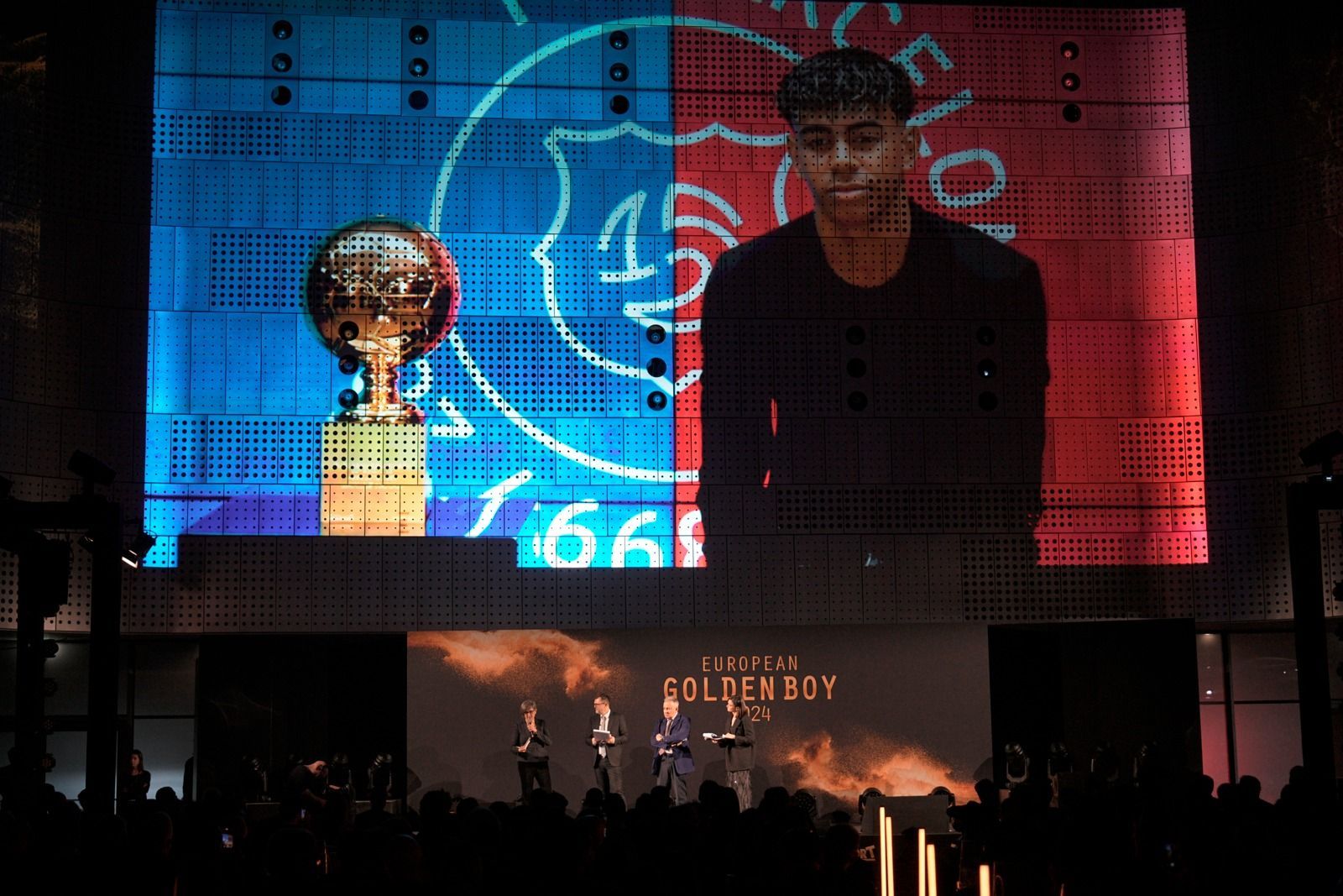 Cerimonia di premiazione del Golden Boy: un trofeo e un ragazzo vengono mostrati su uno schermo; i presentatori parlano su un palco.