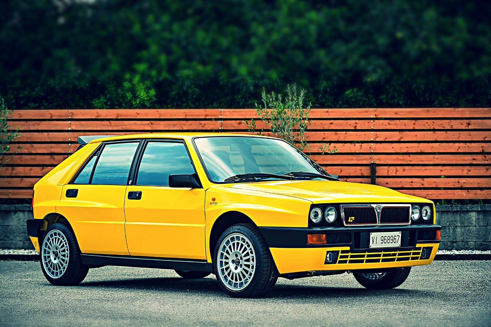 Un'auto da rally Lancia Delta Integrale gialla parcheggiata davanti a un muro con listelli di legno.