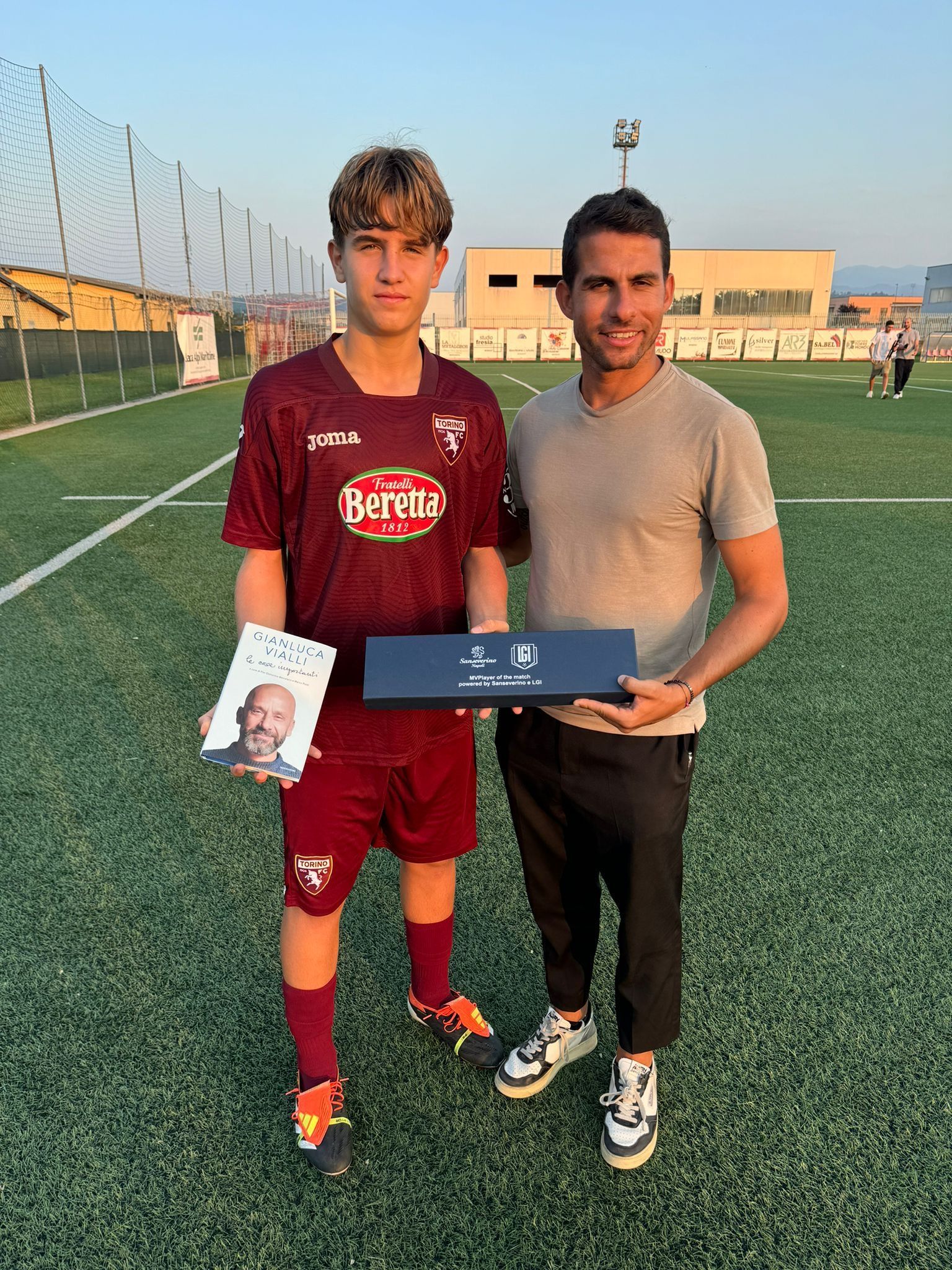 Un giovane calciatore in uniforme marrone e un uomo che regge un trofeo su un campo verde.