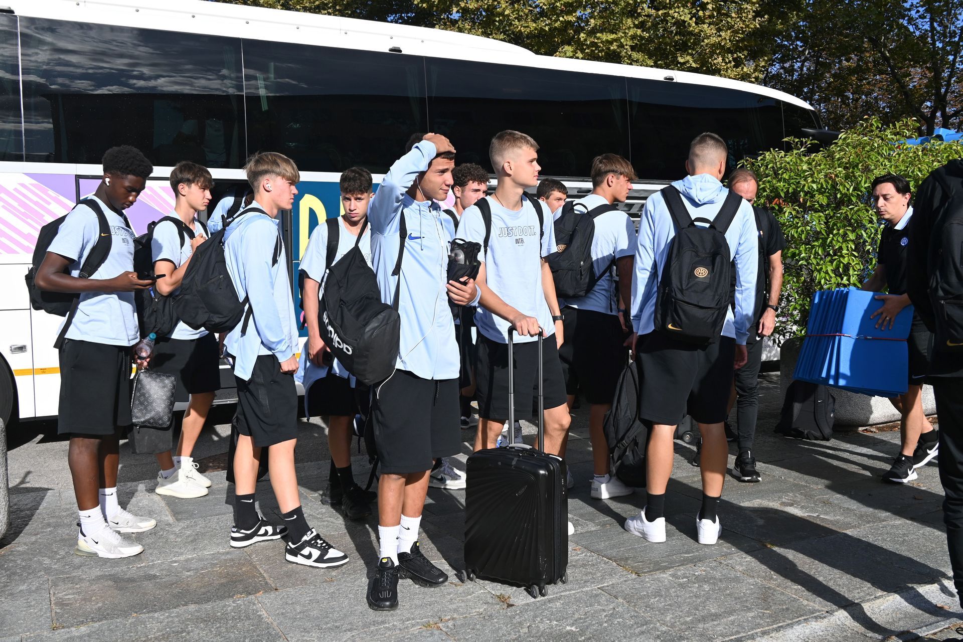 Squadra di calcio, in maglietta azzurra e pantaloncini neri, mentre scende da un autobus. Bagagli visibili. Giornata di sole.