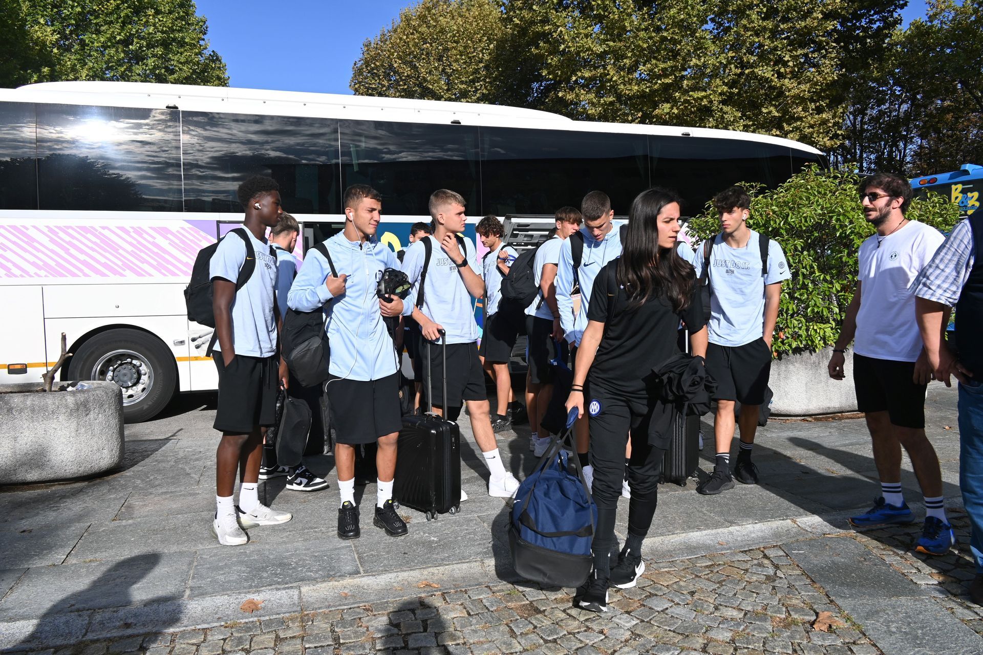 Un gruppo di persone, tra cui alcune con indosso magliette azzurre, in piedi vicino a un autobus. All'aperto, giornata di sole.