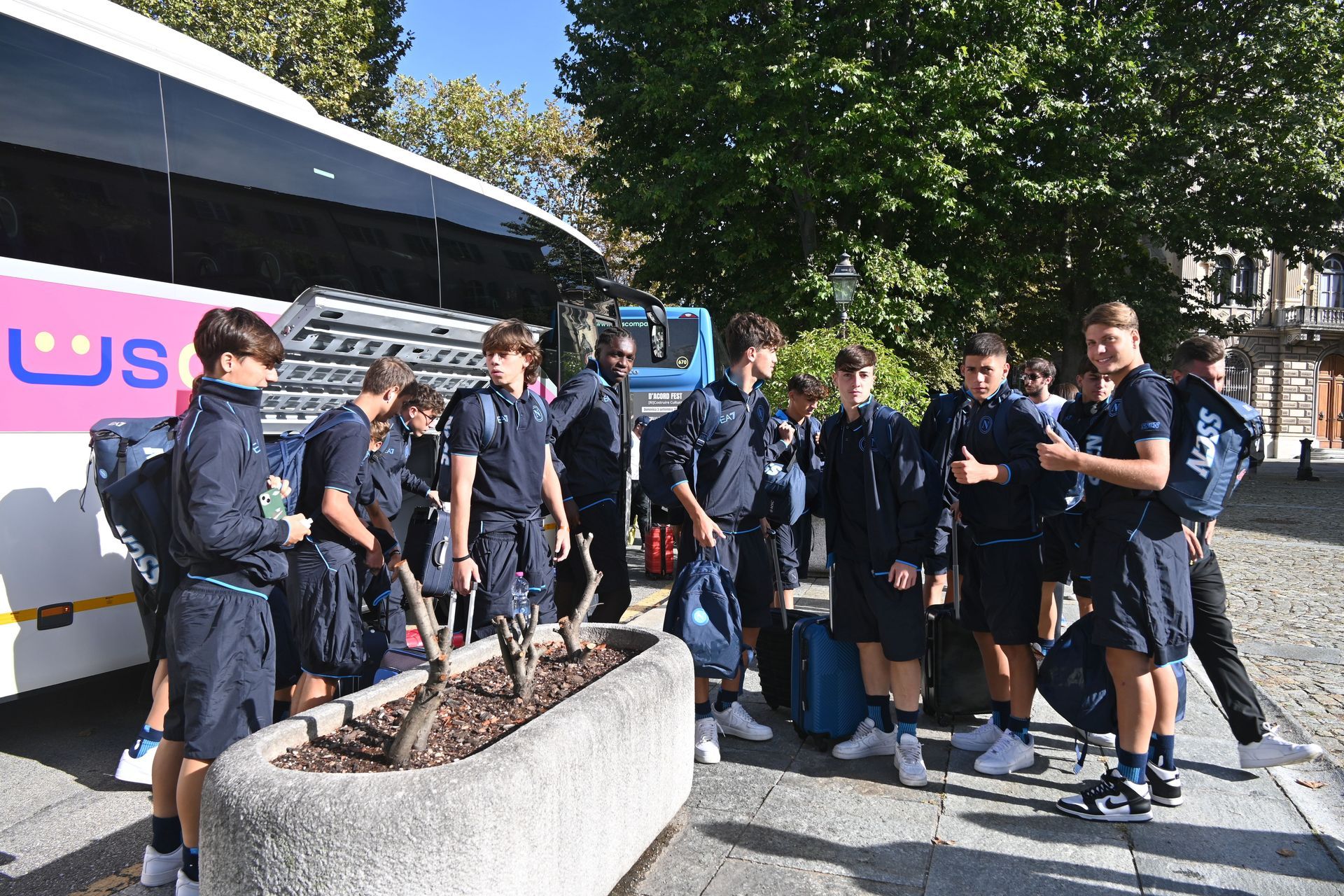 Gruppo di persone in abiti blu coordinati che scendono da un autobus. Bagagli nelle vicinanze. All'aperto, giornata di sole.