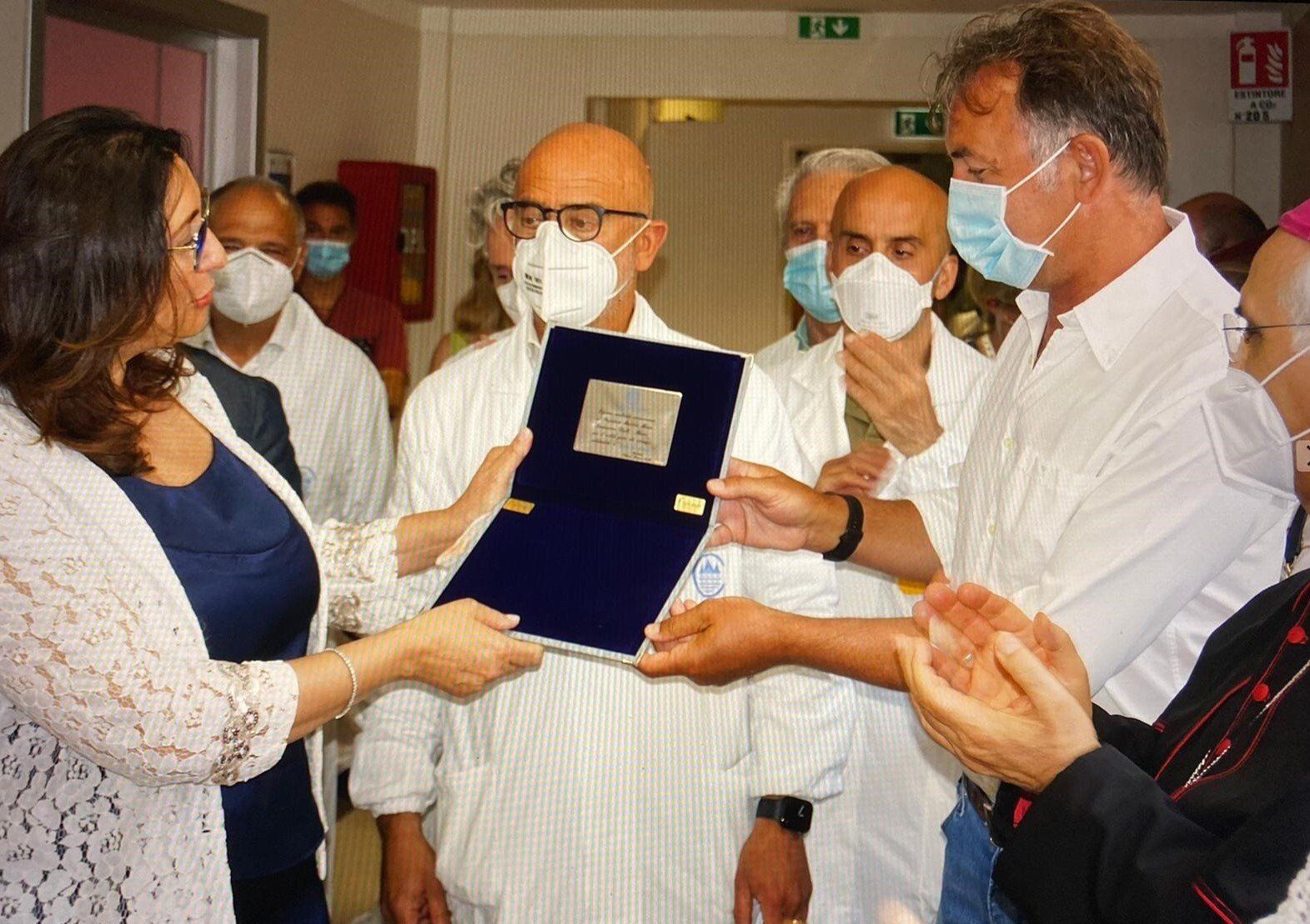 Una donna porge una targa a un uomo, mentre altri osservano. Ambiente interno, camicie bianche, alcune mascherine.